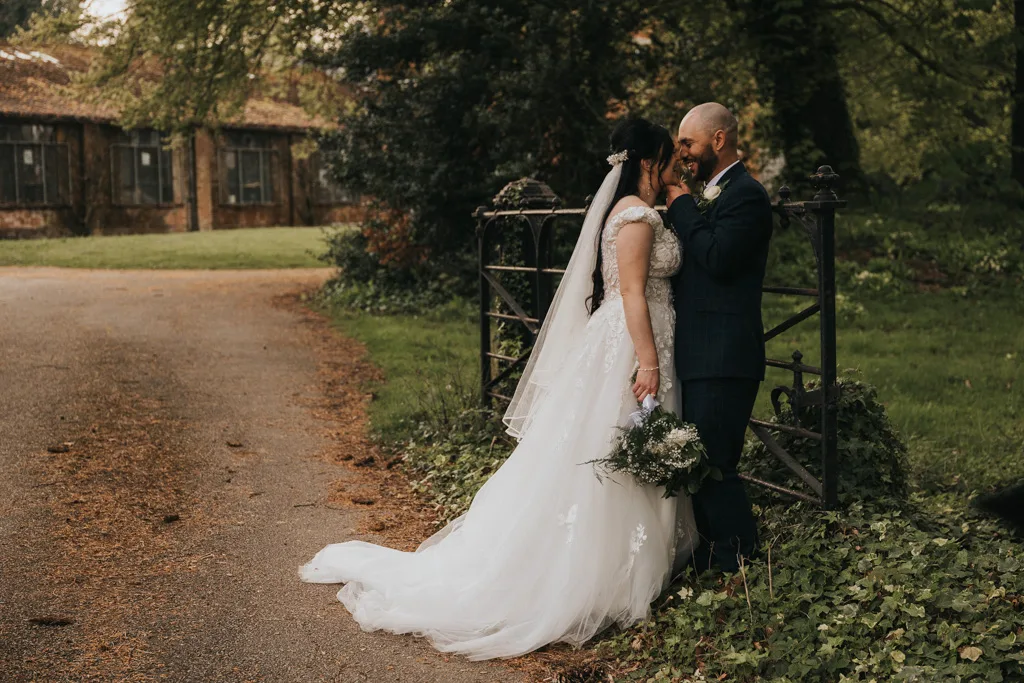 A bride in a white gown and veil holding a bouquet stands closely with her groom by a rustic iron fence, sharing a joyful moment. Perfect for a wedding portfolio, this image captures genuine love amid timeless scenery. © Aimee Lince Photography - Wedding photographer in Lincolnshire, Yorkshire & Nottinghamshire