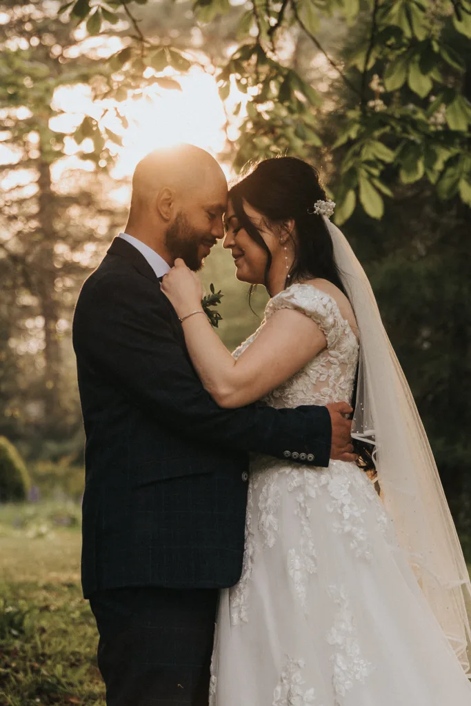 A bride and groom embrace outdoors at sunset, a romantic moment perfect for any portfolio. They touch foreheads, eyes closed and smiling softly as sunlight filters through trees, casting a golden glow over the lace gown and dark suit. © Aimee Lince Photography - Wedding photographer in Lincolnshire, Yorkshire & Nottinghamshire