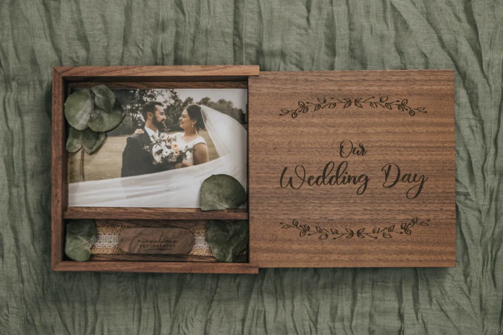 A wooden box with “Our Wedding Day” engraved on the lid sits on green fabric—a perfect keepsake for Yorkshire wedding photography, containing a bride and groom photo, green leaves, lace, and a wooden USB drive labeled “memories to treasure.”. © Aimee Lince Photography - Wedding photographer in Lincolnshire, Yorkshire & Nottinghamshire