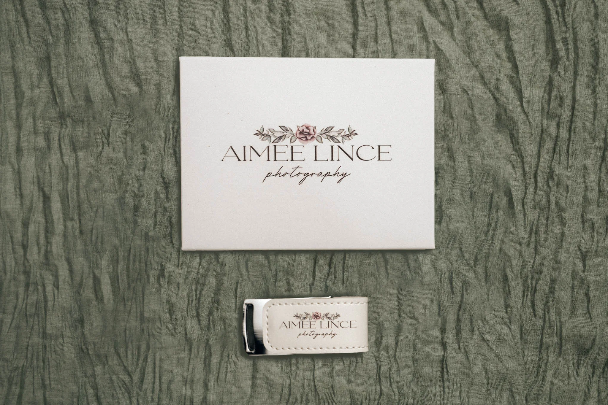 A square white card with "Aimee Lince Photography" and floral graphics sits on wrinkled green fabric. Below the card is a white leather USB drive case with the same logo, closed with visible stitching. © Aimee Lince Photography - Wedding photographer in Lincolnshire, Yorkshire & Nottinghamshire