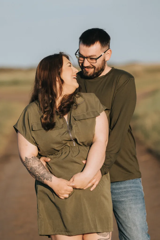 A smiling couple stands on a dirt path in Lincolnshire, embracing. The woman, with long brown hair and a tattooed arm, wears an olive green dress. The man wears glasses and a green shirt. Their engagement photos capture their affectionate gaze outdoors. © Aimee Lince Photography - Wedding photographer in Lincolnshire, Yorkshire & Nottinghamshire