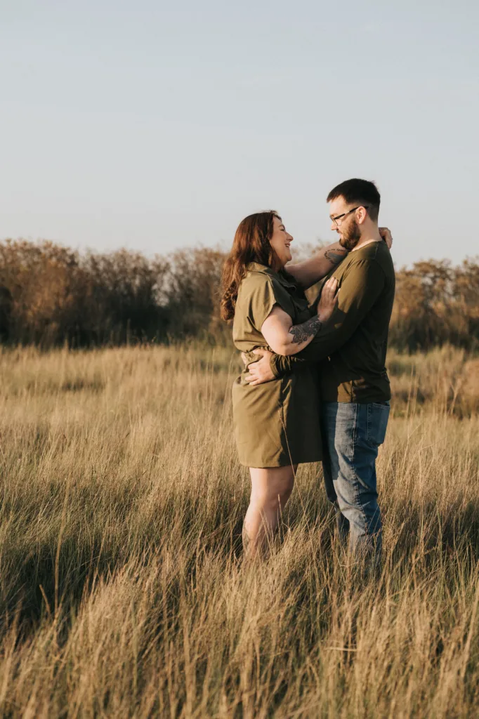 A couple stands in a grassy Cleethorpes field, embracing as they smile warmly at each other. The woman wears an olive dress, the man glasses and a green shirt. Trees and a clear Lincolnshire sky form the perfect backdrop for their photos. © Aimee Lince Photography - Wedding photographer in Lincolnshire, Yorkshire & Nottinghamshire