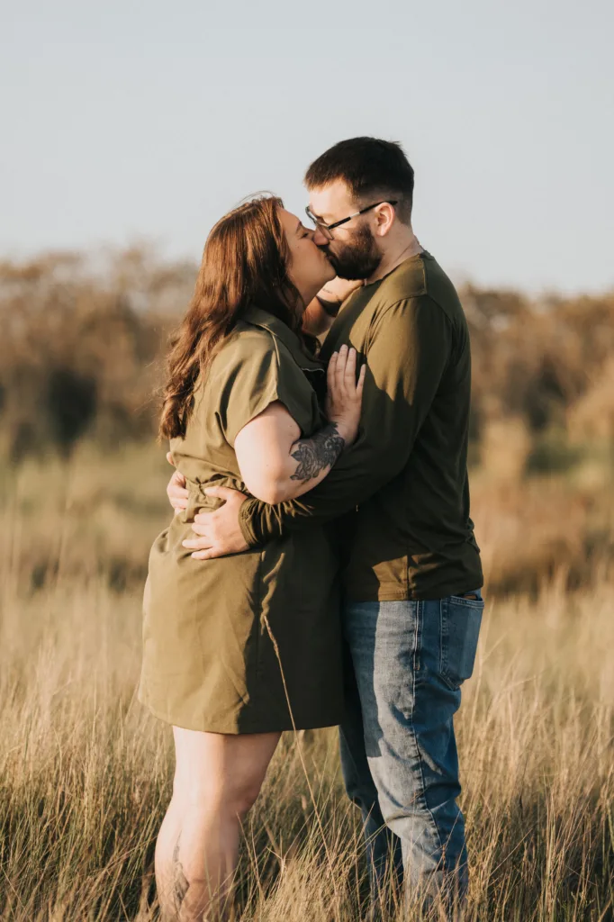 A couple stands in a grassy Lincolnshire field, embracing and sharing a kiss. The woman has long brown hair and a short-sleeved olive green dress, while the man sports a beard, glasses, and jeans. The background of these photos is softly blurred. © Aimee Lince Photography - Wedding photographer in Lincolnshire, Yorkshire & Nottinghamshire