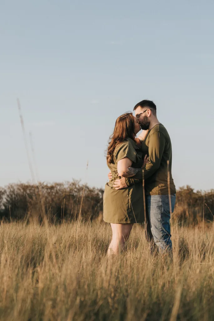 A couple stands in tall grass, embracing and facing each other for photos. The woman wears a short olive dress, and the man wears jeans and an olive shirt. They are outdoors in Cleethorpes, Lincolnshire, with warm sun and distant trees behind them. © Aimee Lince Photography - Wedding photographer in Lincolnshire, Yorkshire & Nottinghamshire