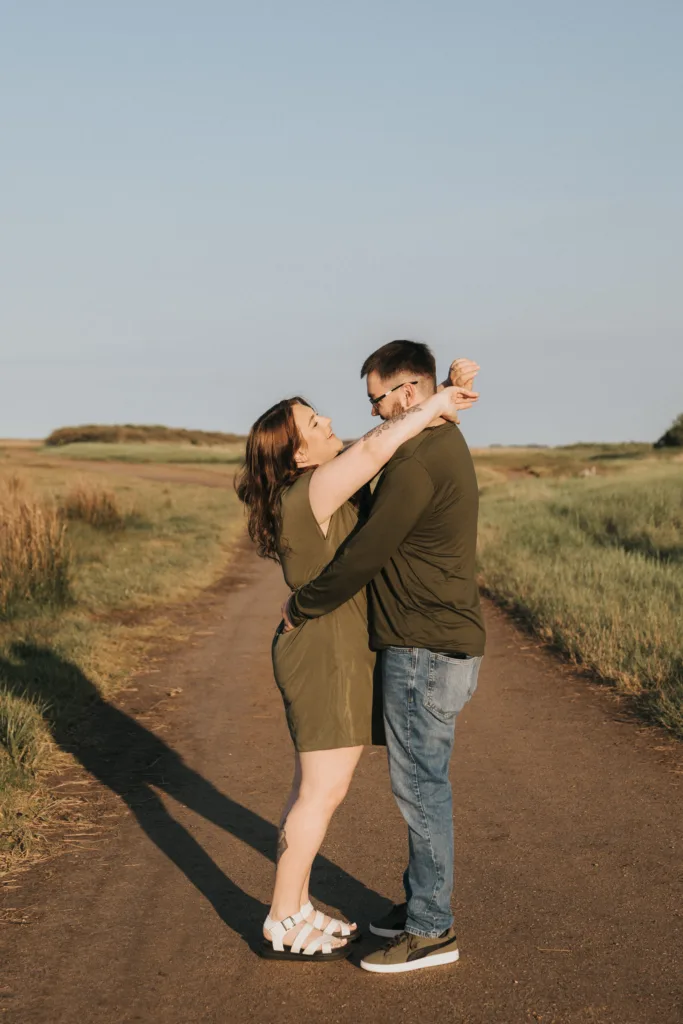A woman in a green dress and white sandals smiles with her arms raised as she stands on a rural path near Cleethorpes, facing a man in jeans and a green shirt who holds her waist. The sun is low, casting long shadows—perfect for engagement photos. © Aimee Lince Photography - Wedding photographer in Lincolnshire, Yorkshire & Nottinghamshire