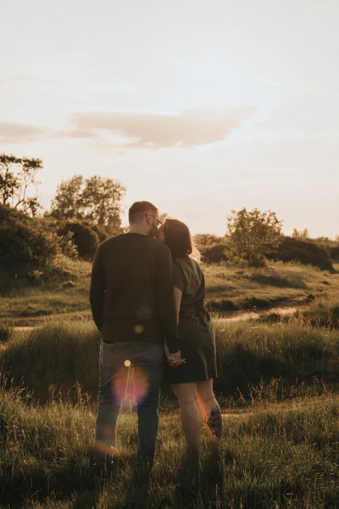 A couple stands hand in hand in a sunlit grassy field at sunset near Cleethorpes, leaning in for a kiss amidst golden light. The sky is partly cloudy with lens flare at their feet—capturing the magic of Lincolnshire photos. © Aimee Lince Photography - Wedding photographer in Lincolnshire, Yorkshire & Nottinghamshire