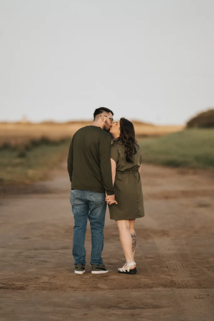 A couple stands on a dirt path in Lincolnshire, holding hands and turning to kiss. The man wears glasses, a green long-sleeve shirt, jeans, and sneakers. The woman wears an olive dress and sandals amid grassy fields beneath a soft, cloudy sky. © Aimee Lince Photography - Wedding photographer in Lincolnshire, Yorkshire & Nottinghamshire