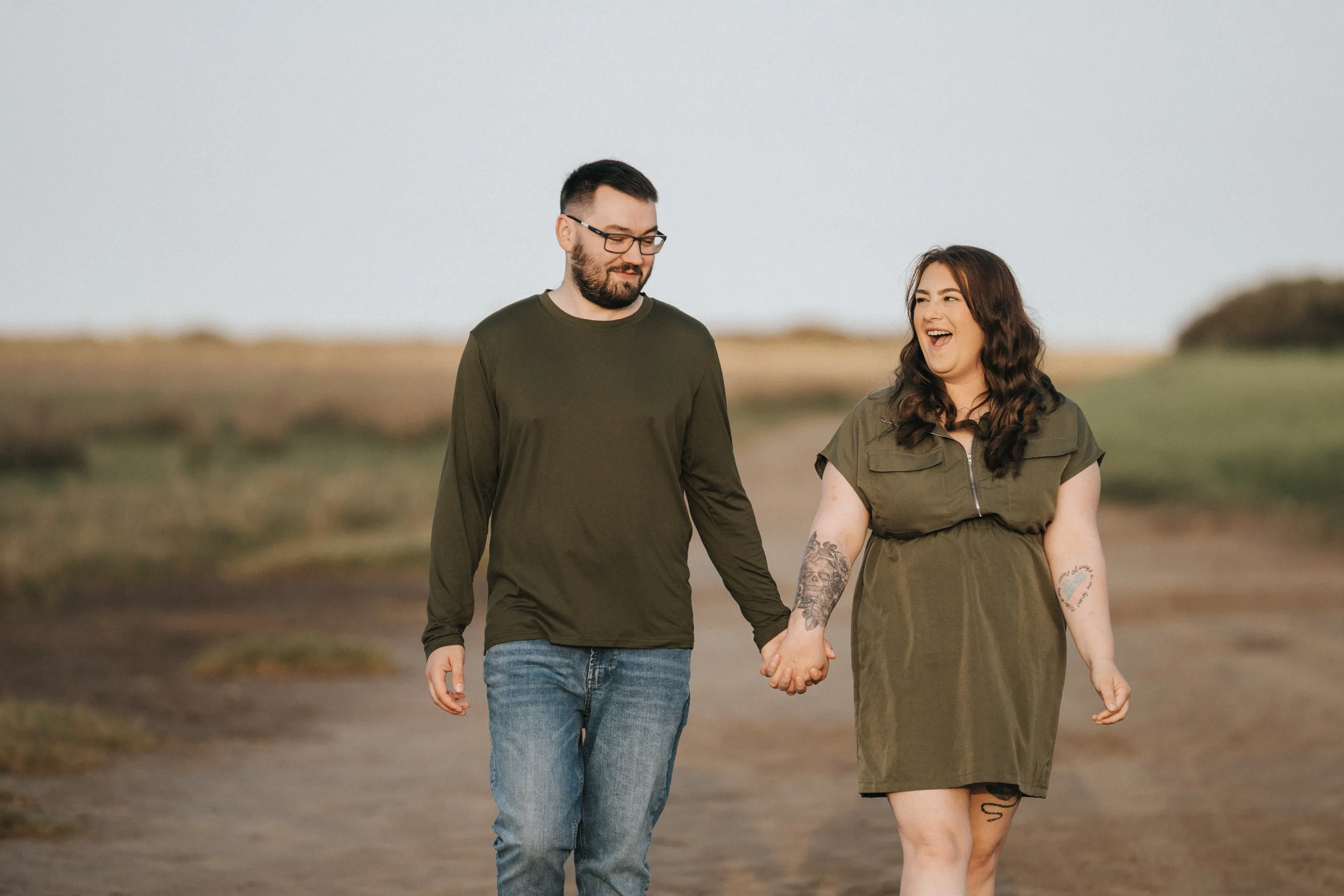 deni-lewis-engagement-photos-cleethorpes-boating-lake-45-of-64-©-aimee-lince-photography-10-06-2025 A man and woman holding hands, walking outdoors on a dirt path in Lincolnshire. Both are smiling; the man wears glasses, a green long-sleeve shirt, and jeans, while the woman wears a short-sleeve green dress and has tattoos on her arms. © Aimee Lince Photography - Wedding photographer in Lincolnshire, Yorkshire & Nottinghamshire