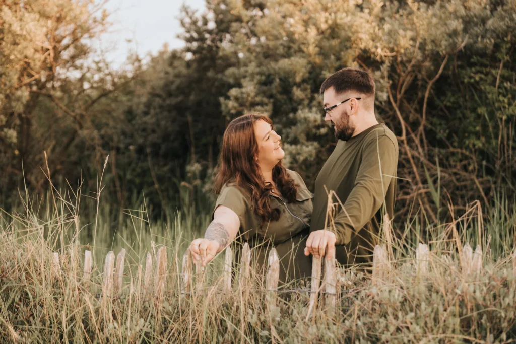 A couple stands in a field of tall grass and wild plants, facing each other and smiling during their engagement photos in Cleethorpes. Both wear olive-green shirts, surrounded by sunlit trees and shrubs for a warm, natural outdoor setting. © Aimee Lince Photography - Wedding photographer in Lincolnshire, Yorkshire & Nottinghamshire