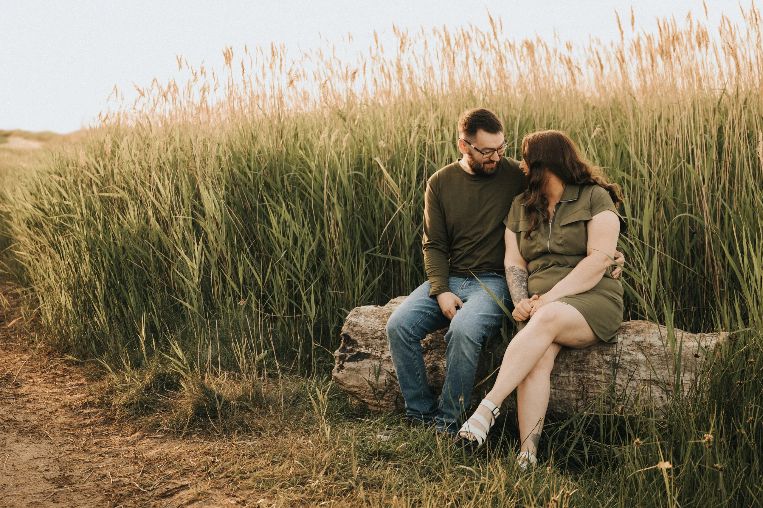 A man and a woman sit close together on a large log in front of tall grass, sharing smiles in warm natural light. This engagement photo, taken in Lincolnshire, captures their joyful connection as they gaze into each other’s eyes. © Aimee Lince Photography - Wedding photographer in Lincolnshire, Yorkshire & Nottinghamshire