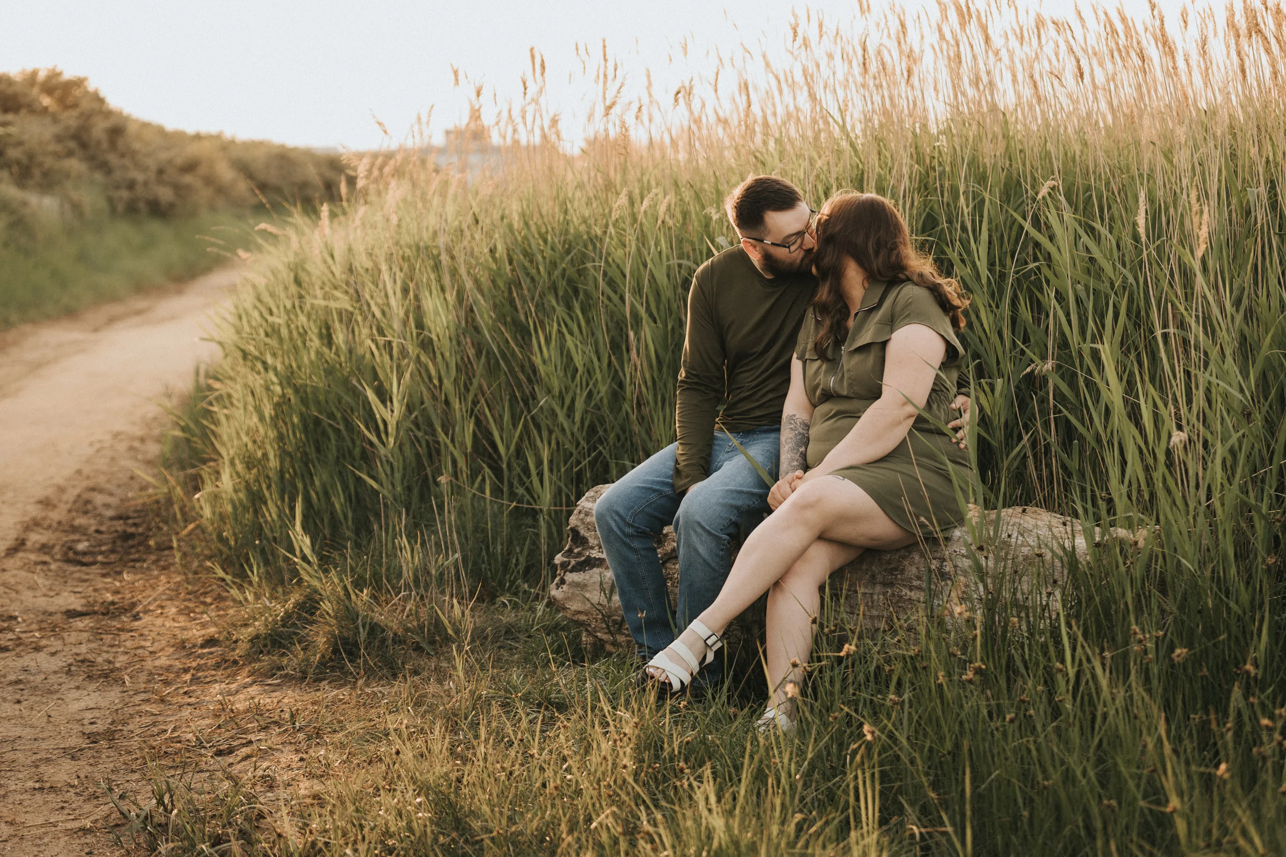 deni-lewis-engagement-photos-cleethorpes-boating-lake-54-of-64-©-aimee-lince-photography-10-06-2025 A couple sits close together on a rock beside a dirt path, surrounded by tall grass. The man wears jeans and a green long-sleeve shirt; the woman wears a short-sleeve olive green dress and white sandals. They lean in for a kiss, sharing an intimate moment at sunset. © Aimee Lince Photography - Wedding photographer in Lincolnshire, Yorkshire & Nottinghamshire