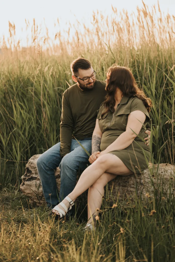 A man and a woman sit close together on a fallen log in a field of tall grass at sunset in Lincolnshire. Sharing warm smiles, this engagement photo captures their joy—the man in glasses and green, the woman in an olive dress and white sandals. © Aimee Lince Photography - Wedding photographer in Lincolnshire, Yorkshire & Nottinghamshire