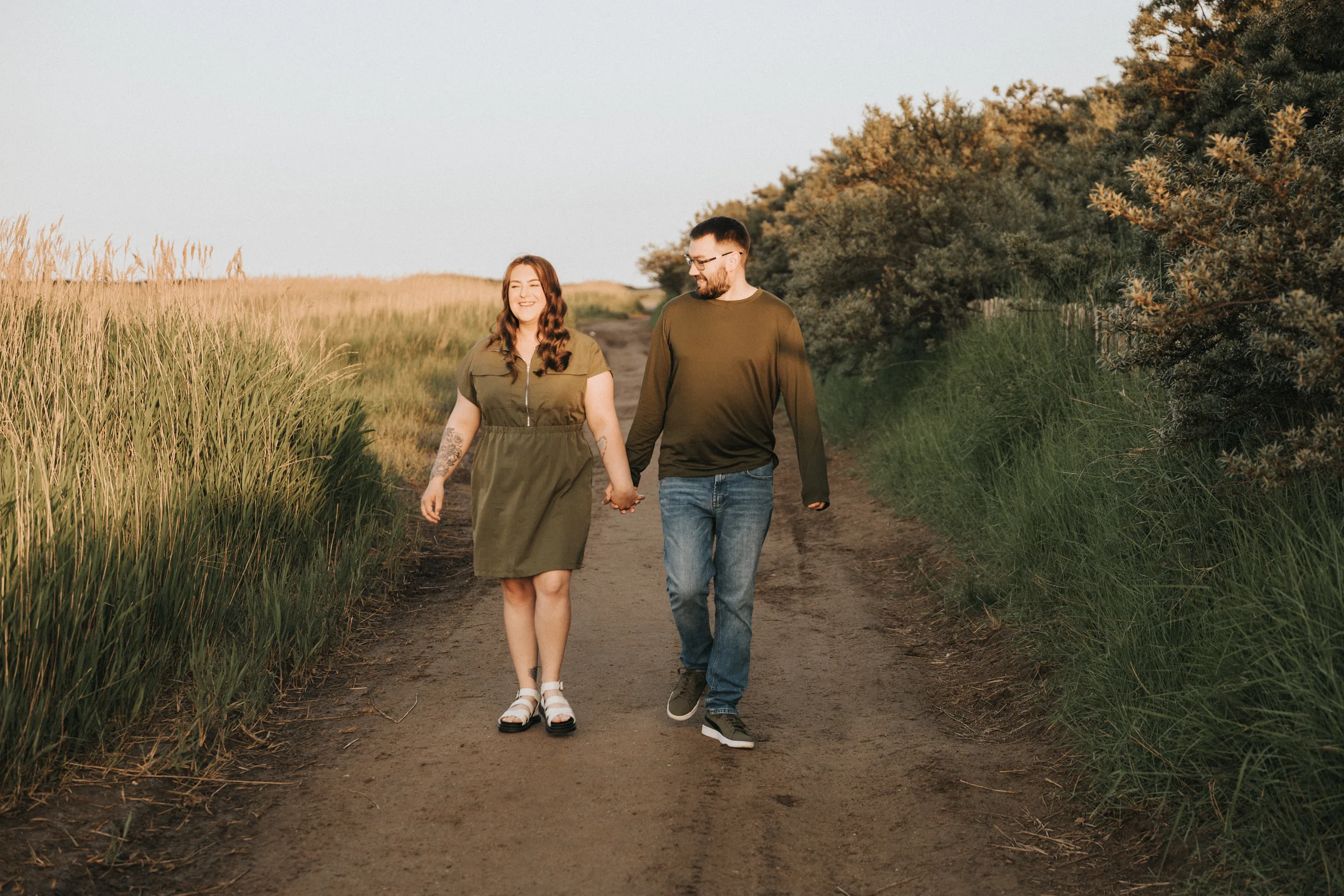 deni-lewis-engagement-photos-cleethorpes-boating-lake-59-of-64-©-aimee-lince-photography-10-06-2025 A man and woman hold hands while walking down a sunlit dirt path surrounded by tall grass in Lincolnshire. Both are smiling and enjoying the natural setting, making it a perfect moment for an engagement stroll. © Aimee Lince Photography - Wedding photographer in Lincolnshire, Yorkshire & Nottinghamshire