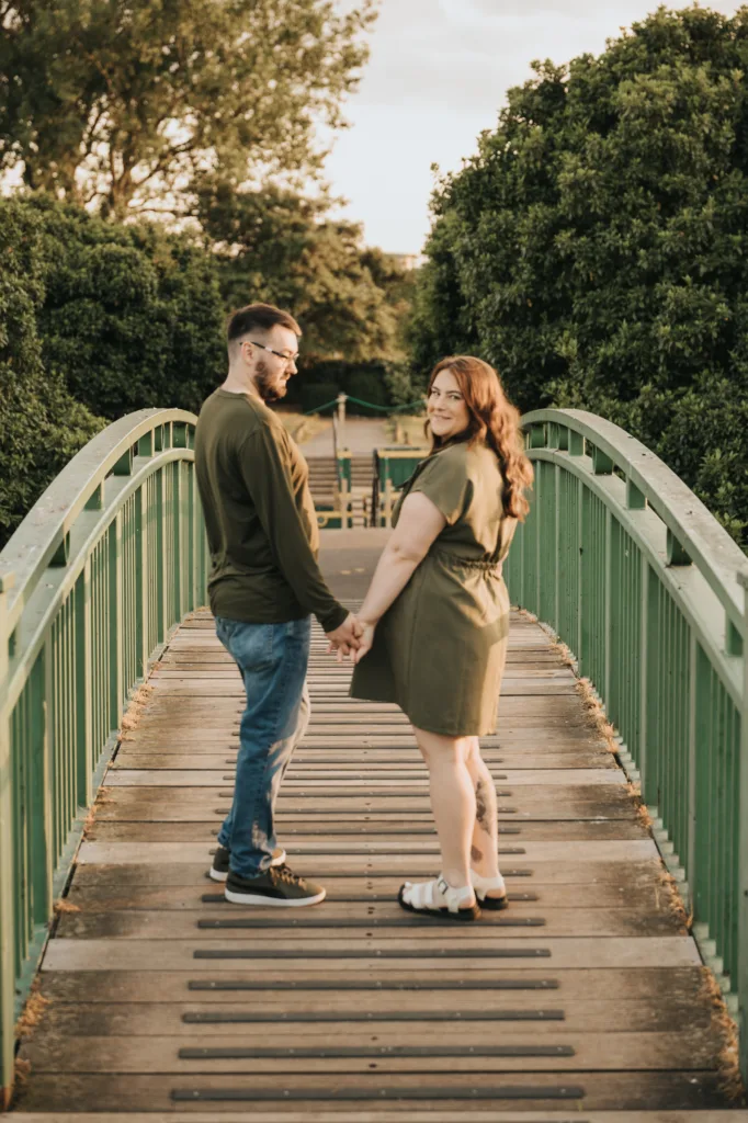 A man and woman stand on a green footbridge in Lincolnshire, holding hands for pre-wedding photos. Both wear green tops; the woman is in a dress and sandals, the man in jeans and sneakers. Sunlight casts a warm, soft glow through the trees. © Aimee Lince Photography - Wedding photographer in Lincolnshire, Yorkshire & Nottinghamshire