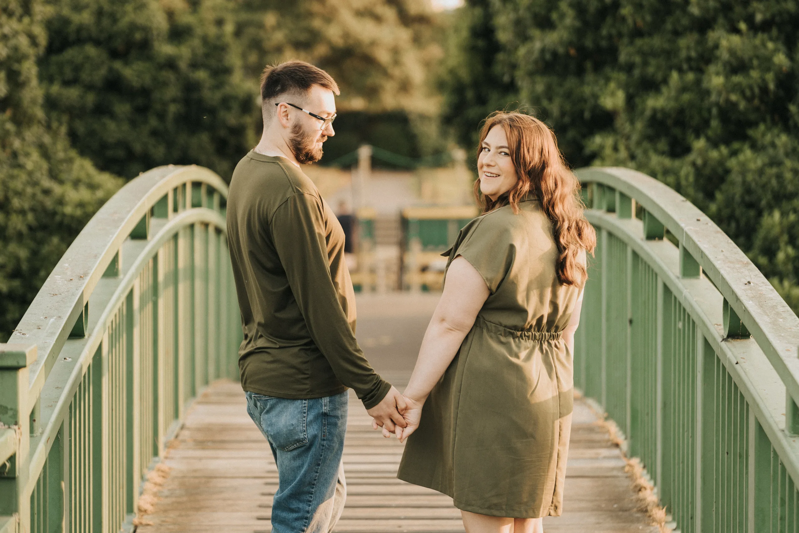 deni-lewis-engagement-photos-cleethorpes-boating-lake-64-of-64-©-aimee-lince-photography-10-06-2025 A couple stands on a green bridge in Lincolnshire holding hands. The man, in a long-sleeve olive shirt and jeans, looks back while the woman, in a short-sleeve olive dress, smiles at the camera—capturing perfect pre-wedding photos amid sunlit trees. © Aimee Lince Photography - Wedding photographer in Lincolnshire, Yorkshire & Nottinghamshire
