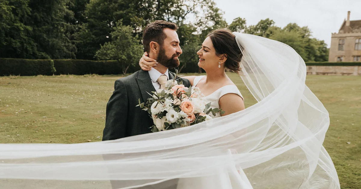 A bride and groom stand outside on a lawn at Rushton Hall, gazing lovingly at each other. The bride’s veil flows in the wind as her bouquet shines—moments beautifully captured by an East Midlands wedding photographer. © Aimee Lince Photography - Wedding photographer in Lincolnshire, Yorkshire & Nottinghamshire