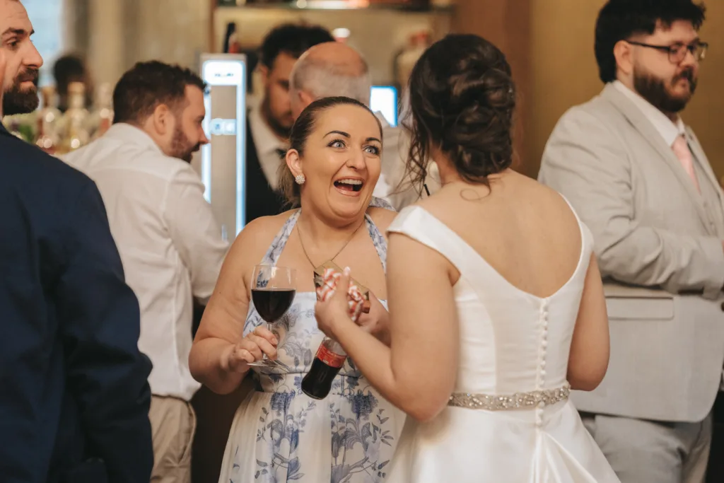 A woman in a blue floral dress, holding a glass of wine and a soda bottle, smiles excitedly while talking to a bride in a white off-shoulder gown at a lively indoor wedding reception. Other guests chat in the warmly lit background. © Aimee Lince Photography - Wedding photographer in Lincolnshire, Yorkshire & Nottinghamshire