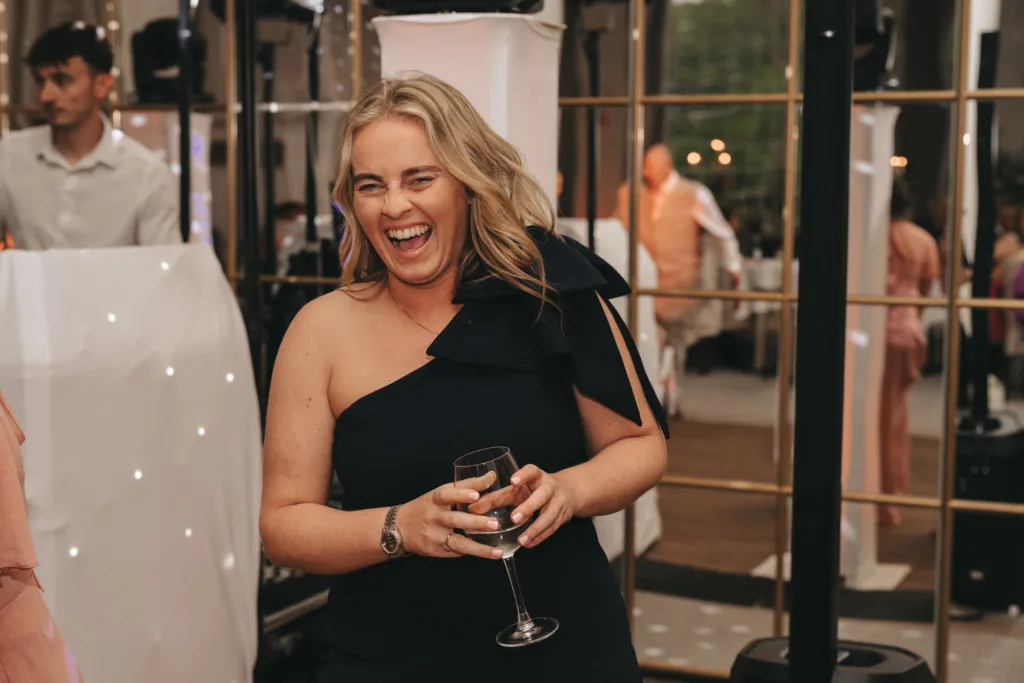 A woman with long blonde hair wearing a one-shoulder black dress laughs joyfully, holding a glass of wine. She stands indoors at a lively event with mirrored panels and other guests visible in the softly lit background. © Aimee Lince Photography - Wedding photographer in Lincolnshire, Yorkshire & Nottinghamshire