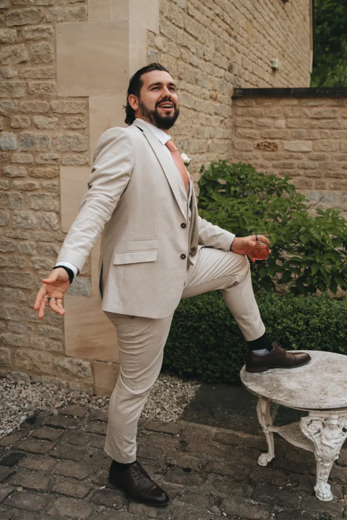 A man with long dark hair and a beard, wearing a light beige suit, white shirt, and peach tie, poses playfully outdoors with one foot on a white metal garden table. He smiles enthusiastically, holding a drink, standing by a stone building and greenery. © Aimee Lince Photography - Wedding photographer in Lincolnshire, Yorkshire & Nottinghamshire