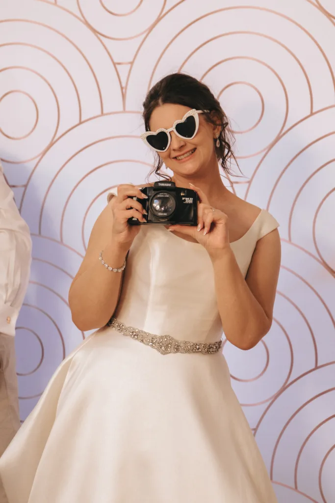 A woman in a white wedding dress and heart-shaped sunglasses smiles while holding a camera. She stands in front of a pastel backdrop with circular patterns, looking cheerful and playful. © Aimee Lince Photography - Wedding photographer in Lincolnshire, Yorkshire & Nottinghamshire