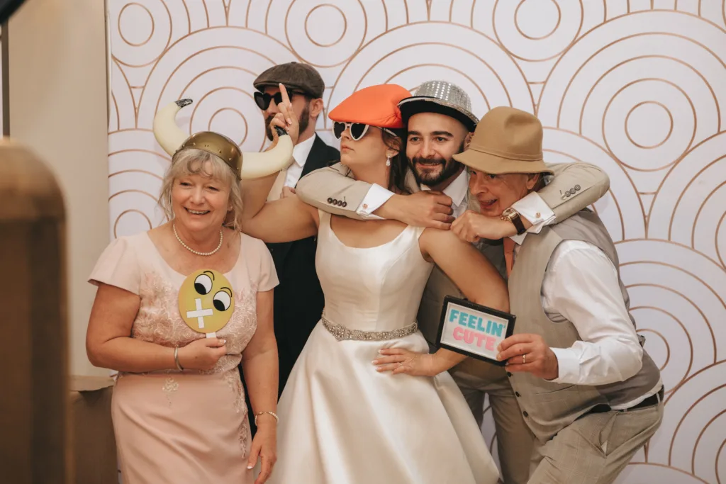 Five people pose playfully in front of a patterned backdrop, wearing fun hats and glasses. The bride stands center in a white dress, flanked by men in suits and hats. One holds a “Feelin’ Cute” sign; another woman smiles holding a funny emoji sign, creating a joyful scene. © Aimee Lince Photography - Wedding photographer in Lincolnshire, Yorkshire & Nottinghamshire