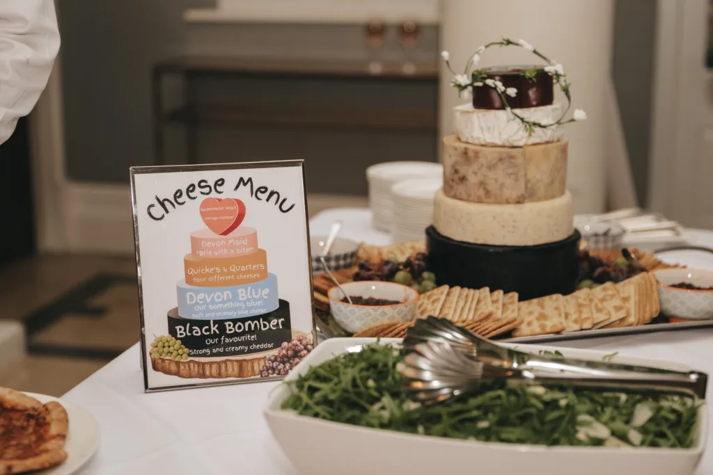 A cheese menu and tiered cheese wheel centerpiece are on a table. The menu lists Devon Place, Quickes Quarters, Devon Blue, and Black Bomber cheeses. Surrounding the cheese are bowls, crackers, grapes, salad, and plates, ready for serving. © Aimee Lince Photography - Wedding photographer in Lincolnshire, Yorkshire & Nottinghamshire