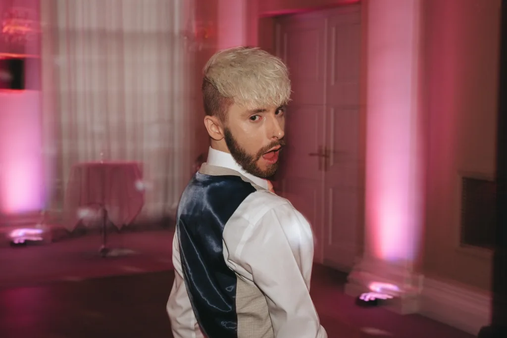 A person with short, platinum blonde hair and a trimmed beard wears a white shirt and dark blue vest, standing indoors with pink lighting. They look over their shoulder at the camera with a playful expression. A round table is blurred in the background. © Aimee Lince Photography - Wedding photographer in Lincolnshire, Yorkshire & Nottinghamshire