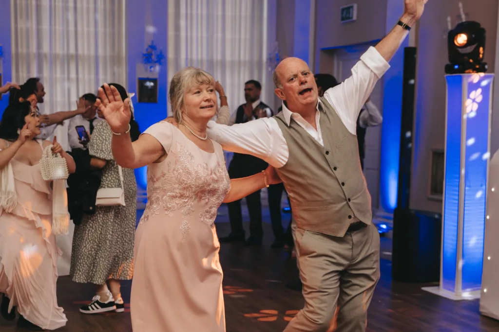 An older couple joyfully dances together at an indoor event. Both have their arms raised and are smiling. The woman wears a pink dress with floral embroidery, and the man wears a white shirt with a gray vest and pants. Other guests and blue lighting are visible in the background. © Aimee Lince Photography - Wedding photographer in Lincolnshire, Yorkshire & Nottinghamshire