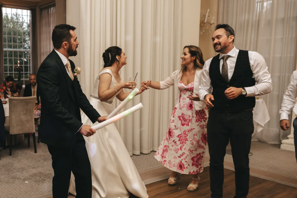 Four adults are dancing and laughing together at an indoor event. A bride in a white dress holds a drink, while a woman in a pink floral dress smiles at her. Two men, one in a dark suit and one in a vest, stand nearby, holding illuminated sticks. © Aimee Lince Photography - Wedding photographer in Lincolnshire, Yorkshire & Nottinghamshire