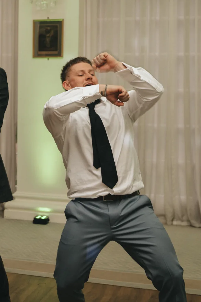 A man in a white dress shirt, untucked black tie, and blue pants energetically dances indoors. His elbows are bent, fists raised, and expression focused. The background features sheer white curtains, a framed picture, and soft, ambient lighting. © Aimee Lince Photography - Wedding photographer in Lincolnshire, Yorkshire & Nottinghamshire
