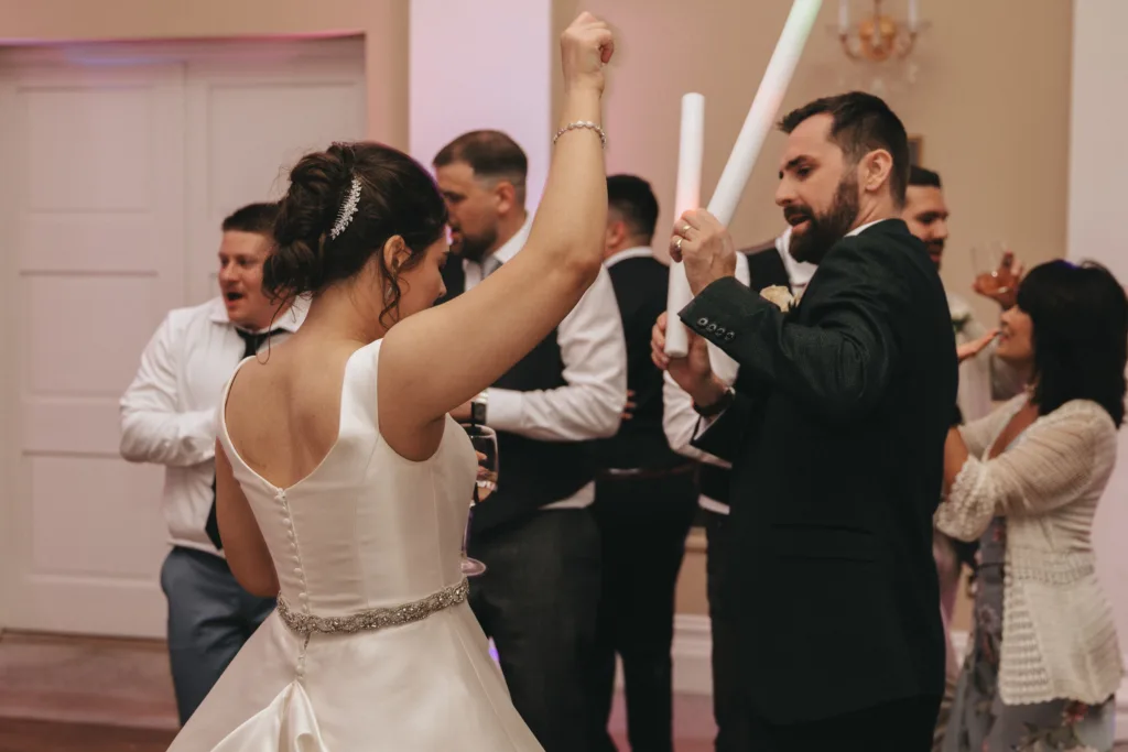 A bride in a white dress dances with guests at a wedding reception. She faces away, raising an arm joyfully. Around her, people in formal wear, including a man with a beard holding a glow stick, join in the lively celebration. © Aimee Lince Photography - Wedding photographer in Lincolnshire, Yorkshire & Nottinghamshire
