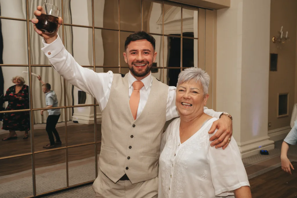 A smiling man in a beige vest and peach tie stands with his arm around an older woman in a white blouse. He raises a drink, celebrating. They pose in front of mirrored panels; reflections and other people are visible in the background. © Aimee Lince Photography - Wedding photographer in Lincolnshire, Yorkshire & Nottinghamshire