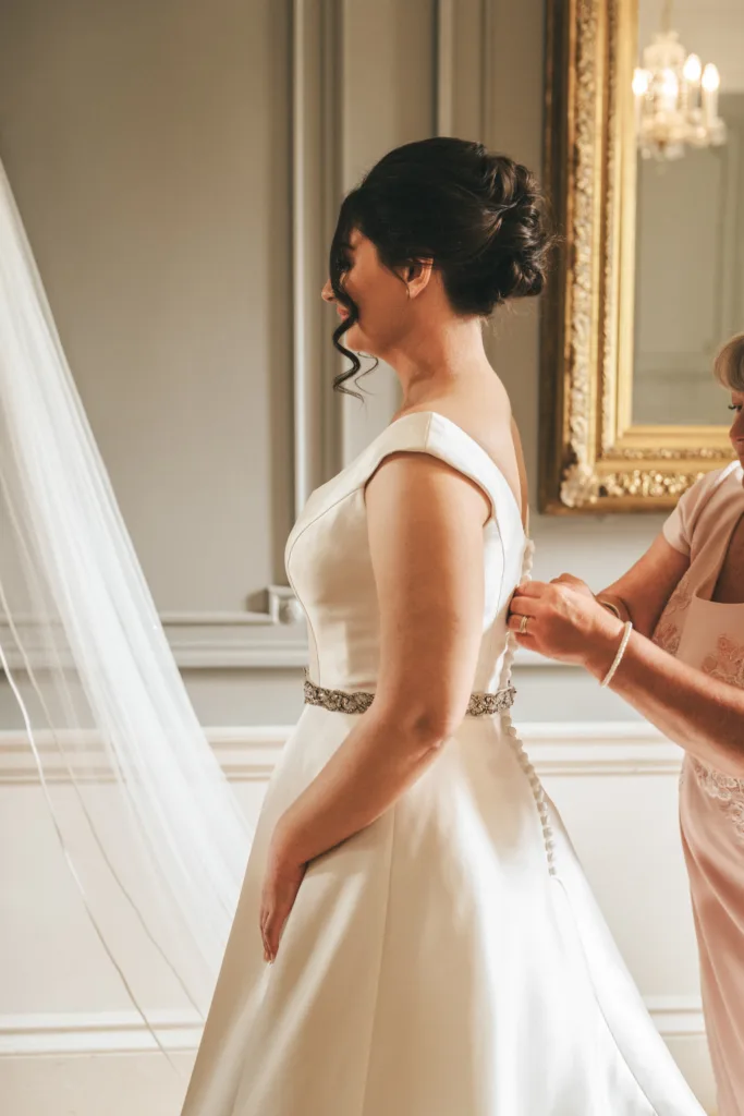 A bride in a white dress stands in profile as an older woman fastens her gown—an intimate moment captured by a candid wedding photographer. The elegant gray room features white trim and a grand gold-framed mirror, reflecting wedding day joy. © Aimee Lince Photography - Wedding photographer in Lincolnshire, Yorkshire & Nottinghamshire