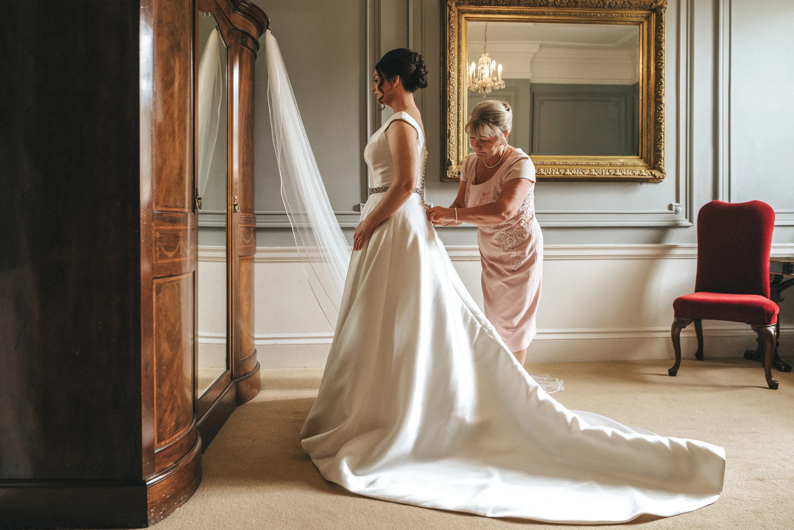 katie-adair-wedding-photography-rushton-hall-kettering-northamptonshire-120-of-1141-©-aimee-lince-photography-02-06-2025 A bride in a white gown stands by a wardrobe as an older woman fastens her dress. Elegant decor fills the room at this Rushton Hall wedding: a gold-framed mirror, gray walls, chandelier, red chair, and the bride’s long train flowing onto the floor. © Aimee Lince Photography - Wedding photographer in Lincolnshire, Yorkshire & Nottinghamshire
