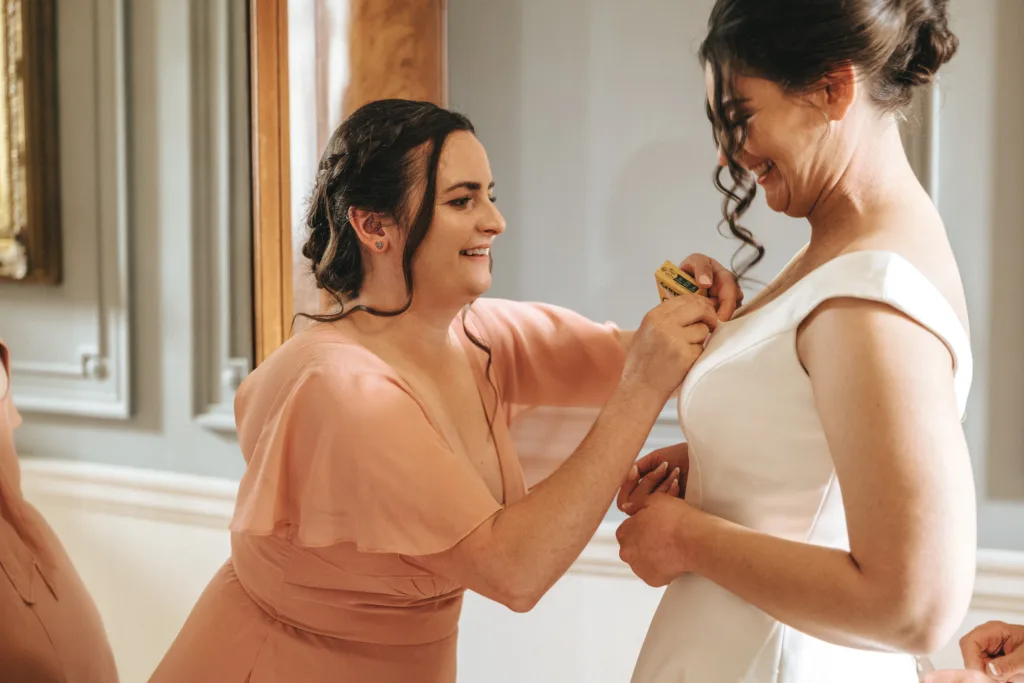 Two women stand indoors in front of ornate paneling. One, in a peach dress, smiles as she pins a yellow measuring tape to the other's white wedding gown. This candid wedding photography moment captures joy, perfect for a Rushton Hall wedding. © Aimee Lince Photography - Wedding photographer in Lincolnshire, Yorkshire & Nottinghamshire