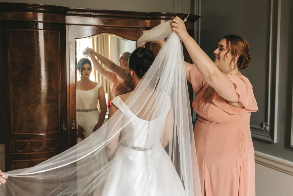 A woman in a peach dress helps a bride adjust her long white veil in front of a mirrored wardrobe. Captured with candid wedding photography, both the bride and bridesmaid are focused on the task, reflecting genuine moments perfect for an East Midlands wedding photographer. © Aimee Lince Photography - Wedding photographer in Lincolnshire, Yorkshire & Nottinghamshire