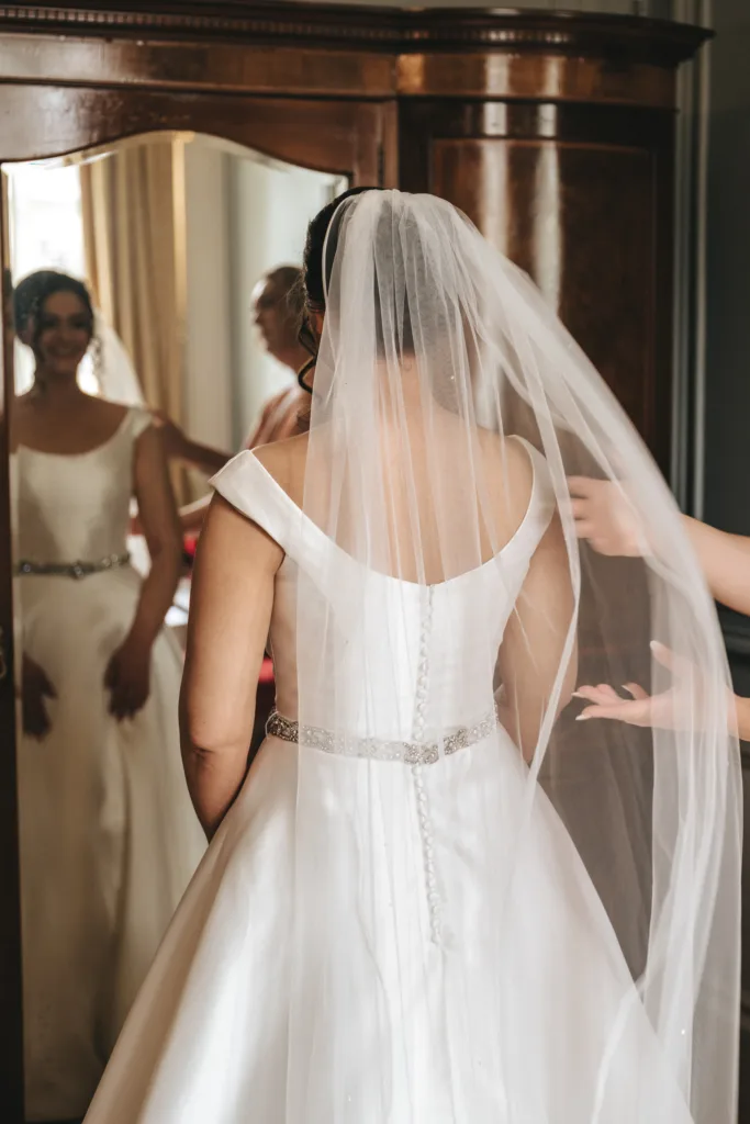 A bride in an off-shoulder white gown with a jeweled belt stands facing away, her veil being adjusted—a candid wedding photography moment. Her smile is reflected in a mirror on a wooden wardrobe behind her. © Aimee Lince Photography - Wedding photographer in Lincolnshire, Yorkshire & Nottinghamshire