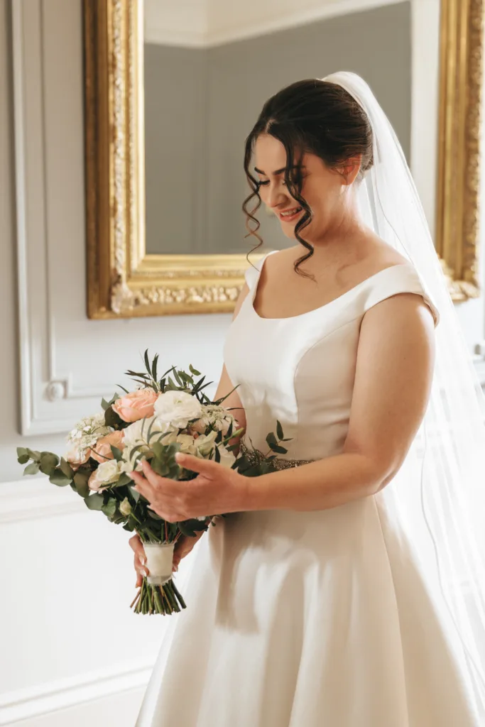 A smiling bride in a white dress and veil holds a bouquet of pale pink and white flowers with greenery. Captured with candid wedding photography at Rushton Hall, she stands near an ornate gold-framed mirror, bathed in soft light. © Aimee Lince Photography - Wedding photographer in Lincolnshire, Yorkshire & Nottinghamshire