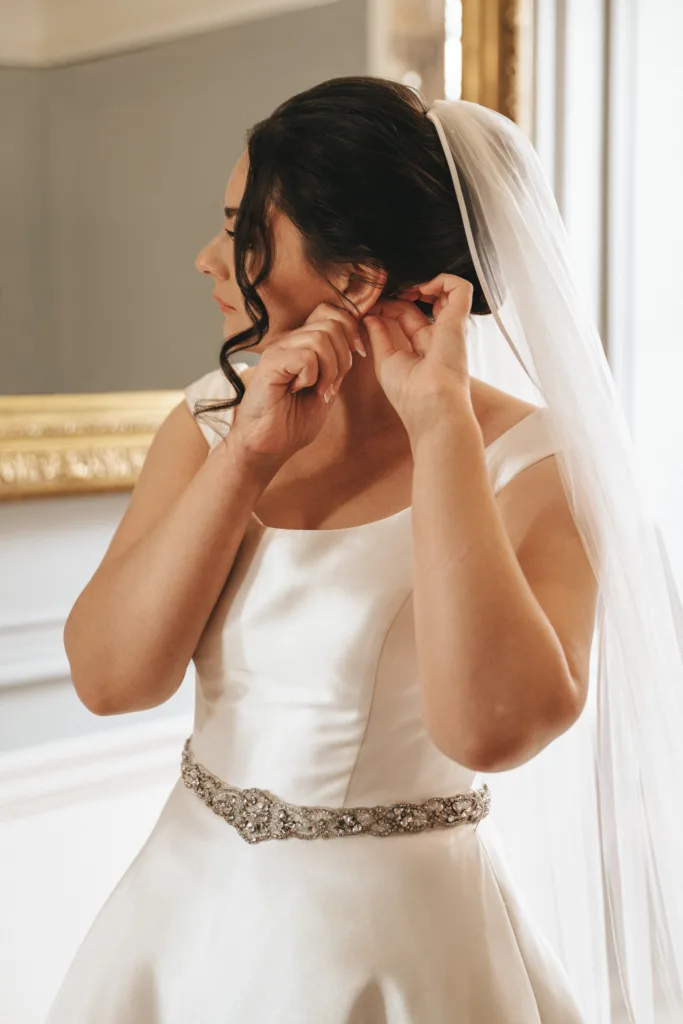 A bride in a white satin gown with a jeweled belt and long veil adjusts an earring. Captured in soft natural light by a Kettering wedding photographer, she stands near a gold-framed mirror—perfect for candid wedding photography at Rushton Hall. © Aimee Lince Photography - Wedding photographer in Lincolnshire, Yorkshire & Nottinghamshire