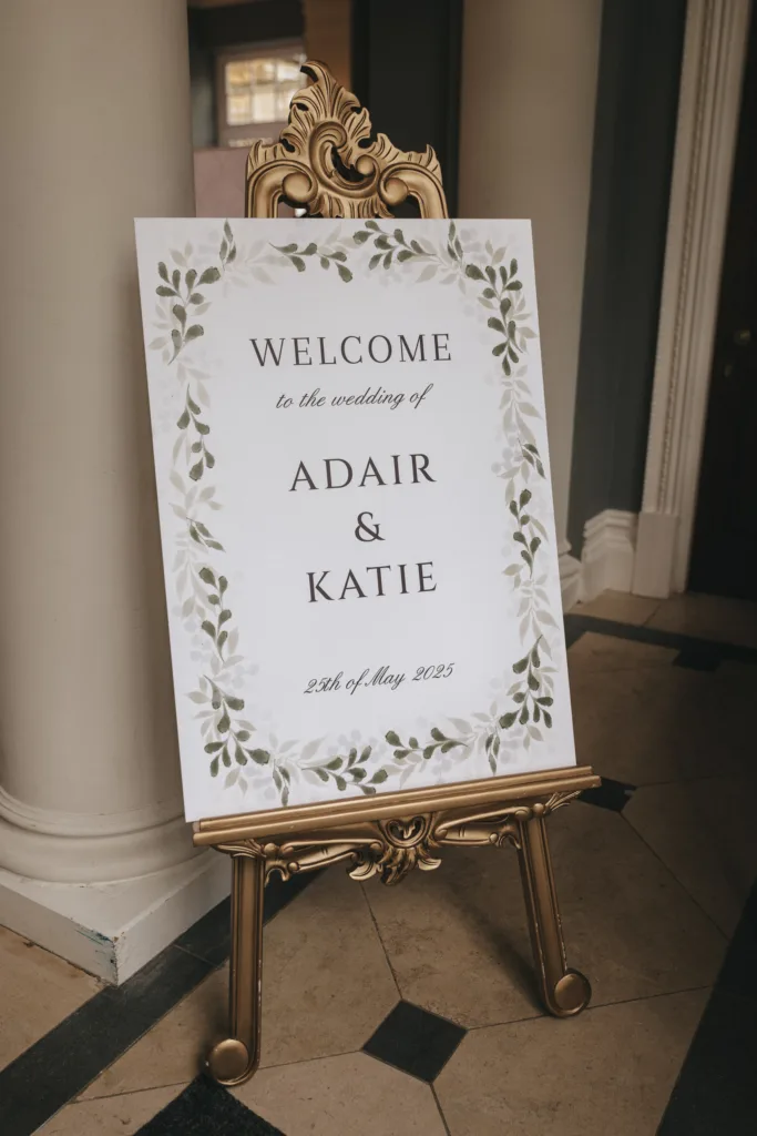 A white wedding welcome sign with olive leaf illustrations reads “Welcome to the wedding of Adair & Katie, 24th of May 2025.” The sign is displayed on an ornate gold easel—perfect inspiration for candid wedding photography. © Aimee Lince Photography - Wedding photographer in Lincolnshire, Yorkshire & Nottinghamshire