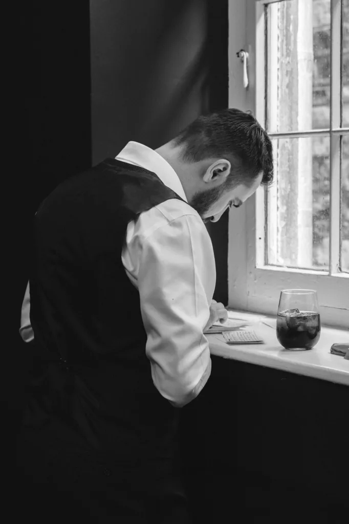 A man in a dress shirt and vest stands by a window at Rushton Hall, leaning over to write on a piece of paper on the windowsill. Next to him is a glass of dark liquid. The black and white scene captures candid wedding photography with natural light. © Aimee Lince Photography - Wedding photographer in Lincolnshire, Yorkshire & Nottinghamshire