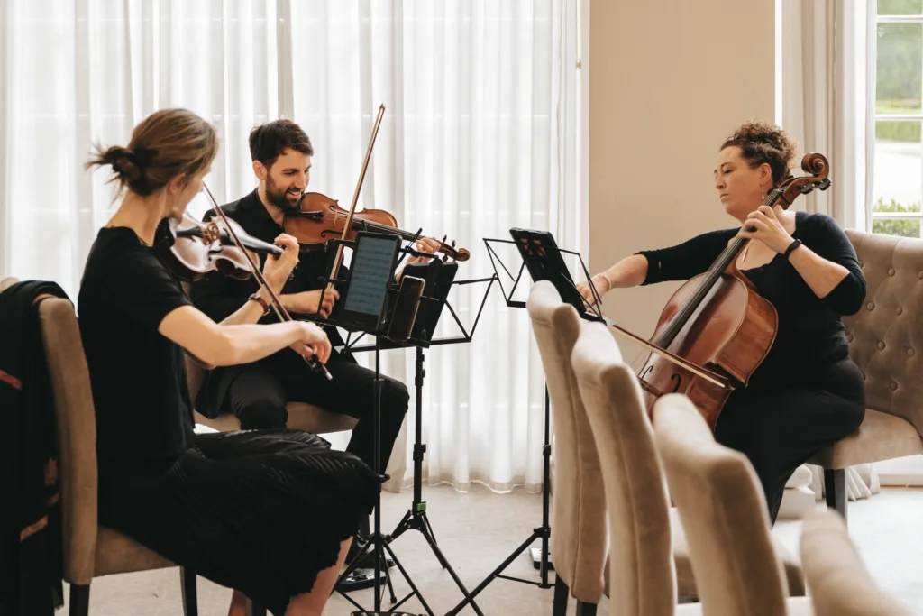 Three musicians sit in a bright room at a Rushton Hall wedding, playing string instruments: two violinists on the left and a cellist on the right. Sunlight filters through sheer white curtains—an elegant moment, perfect for candid wedding photography. © Aimee Lince Photography - Wedding photographer in Lincolnshire, Yorkshire & Nottinghamshire