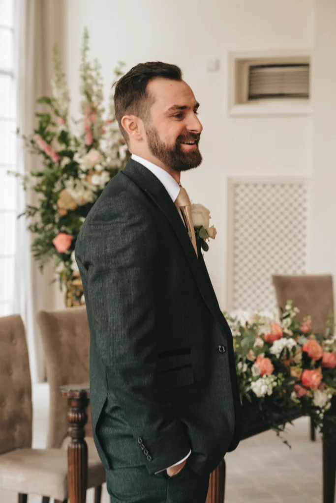 A bearded man in a dark suit with a cream tie and white boutonnière stands smiling indoors at a Rushton Hall wedding. Behind him are beige chairs, lush floral arrangements, and soft natural light from large windows—captured by an East Midlands wedding photographer. © Aimee Lince Photography - Wedding photographer in Lincolnshire, Yorkshire & Nottinghamshire