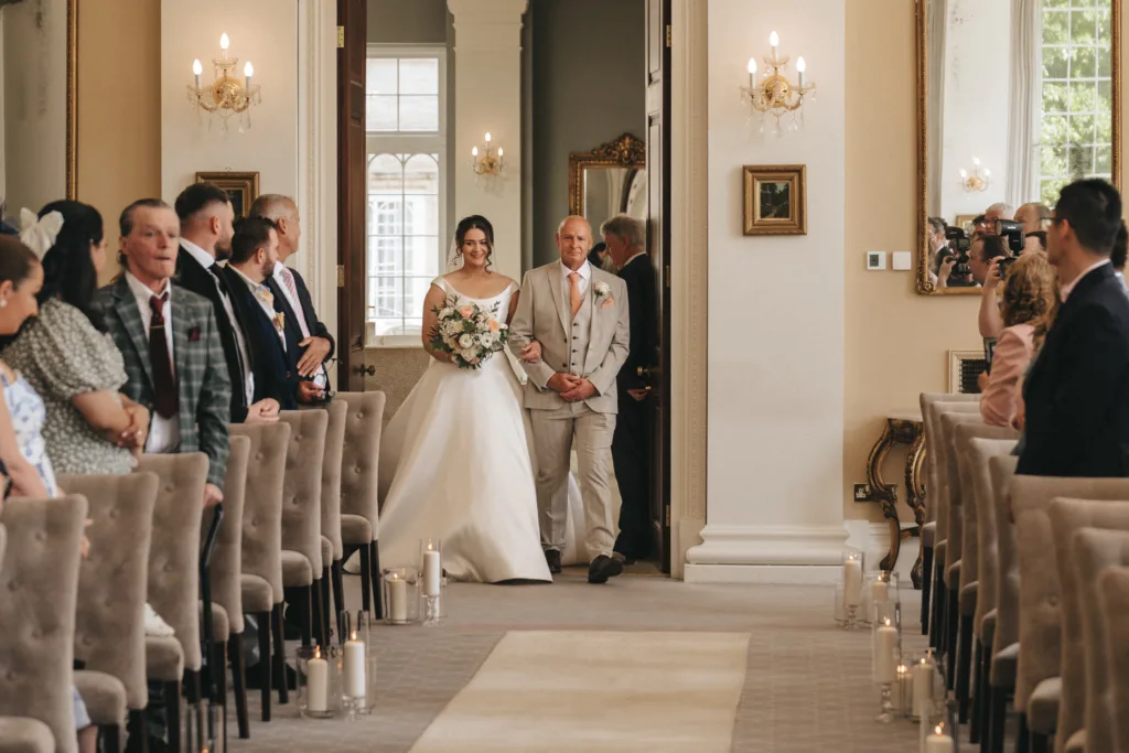 A bride in a white gown walks down the aisle at a Rushton Hall wedding, bouquet in hand and guests smiling. Cream walls, chandeliers, candles, framed art, and daylight from large windows create an elegant setting for this special moment. © Aimee Lince Photography - Wedding photographer in Lincolnshire, Yorkshire & Nottinghamshire