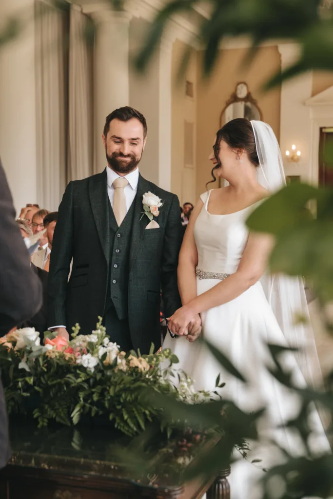 A bride in a white dress and veil stands beside a groom in a dark suit and cream tie. Captured with candid wedding photography, they smile and hold hands during a Rushton Hall wedding, surrounded by greenery, flowers, and guests. © Aimee Lince Photography - Wedding photographer in Lincolnshire, Yorkshire & Nottinghamshire