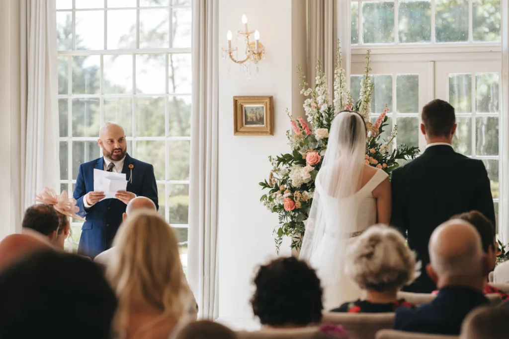 A man in a blue suit reads from papers at a Rushton Hall wedding ceremony indoors. The bride and groom face the officiant as guests watch. Candid wedding photography captures tall floral arrangements in the bright, windowed room. © Aimee Lince Photography - Wedding photographer in Lincolnshire, Yorkshire & Nottinghamshire