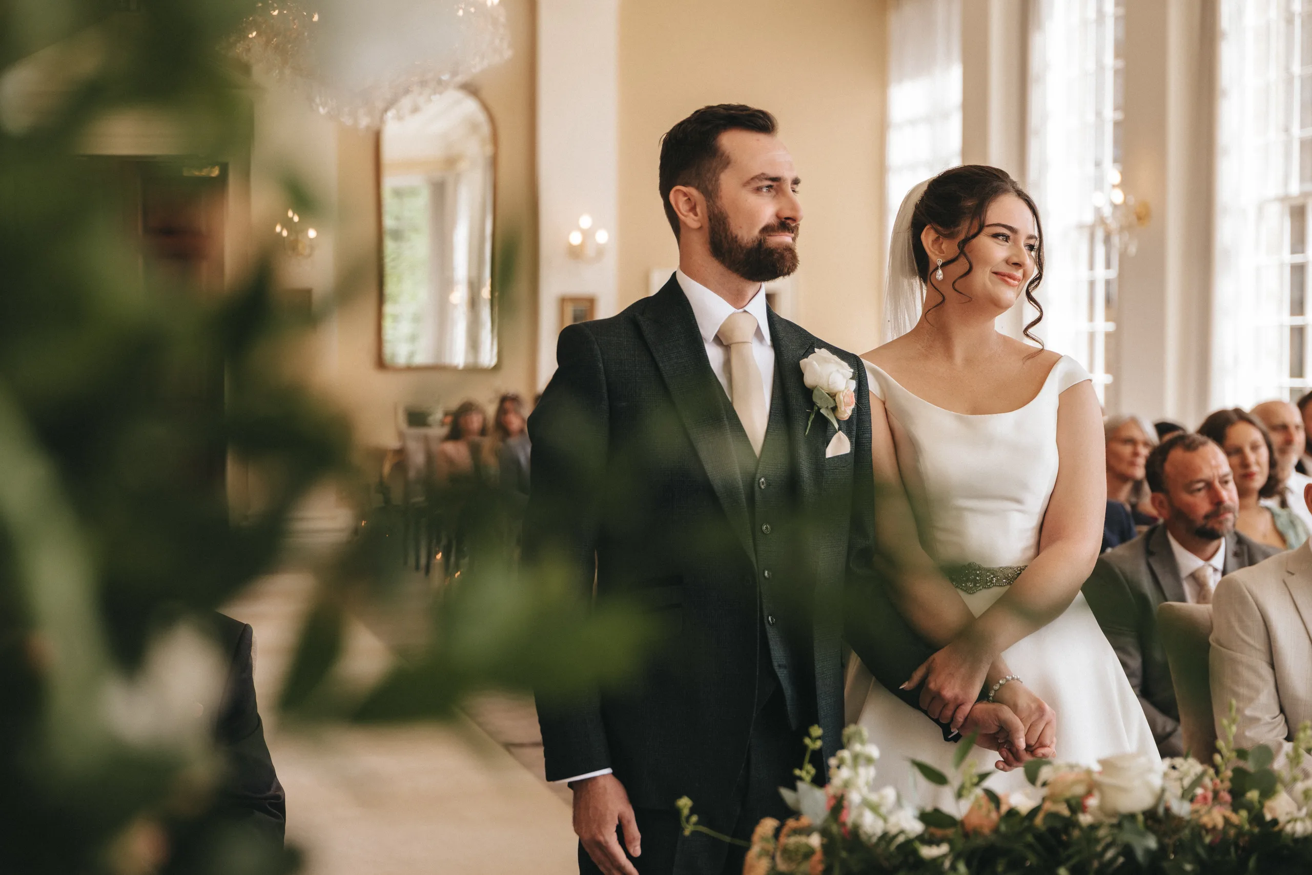 katie-adair-wedding-photography-rushton-hall-kettering-northamptonshire-213-of-1141-©-aimee-lince-photography-02-06-2025 A bride in a white dress and a groom in a dark suit stand side by side at their Rushton Hall wedding, holding hands and smiling. Guests are seated nearby as soft natural light streams through tall windows, with greenery blurred in the foreground. © Aimee Lince Photography - Wedding photographer in Lincolnshire, Yorkshire & Nottinghamshire