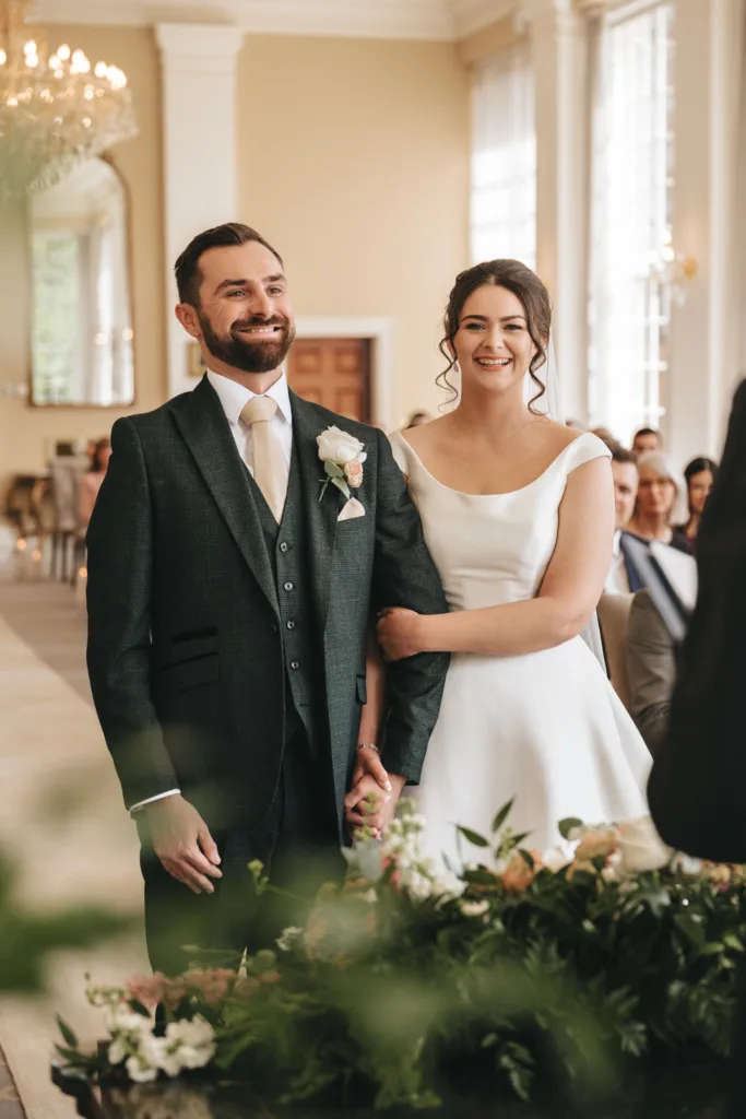 A bride and groom stand smiling at their wedding ceremony, beautifully captured by an east midlands wedding photographer. Guests are seated behind them in a bright, elegant room with large windows, preserving this candid wedding photography moment forever. © Aimee Lince Photography - Wedding photographer in Lincolnshire, Yorkshire & Nottinghamshire