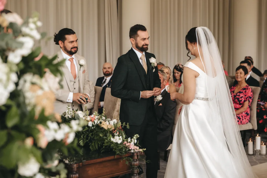 A bride in a white dress and veil stands facing a groom in a dark suit holding a ring. An officiant in beige stands nearby as guests watch. Candid wedding photography captures the floral decor and neutral curtains at this heartfelt ceremony. © Aimee Lince Photography - Wedding photographer in Lincolnshire, Yorkshire & Nottinghamshire