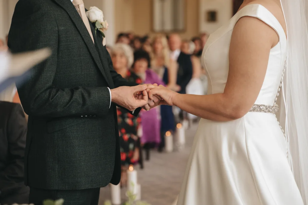 A bride and groom stand facing each other during their Rushton Hall wedding ceremony. The groom places a ring on the bride’s finger as an East Midlands wedding photographer captures the moment. Guests are seated in the blurred background. © Aimee Lince Photography - Wedding photographer in Lincolnshire, Yorkshire & Nottinghamshire